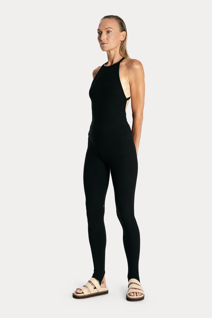 Model wearing a black ribbed bodysuit and matching black stirrup leggings, full-body front-left view.