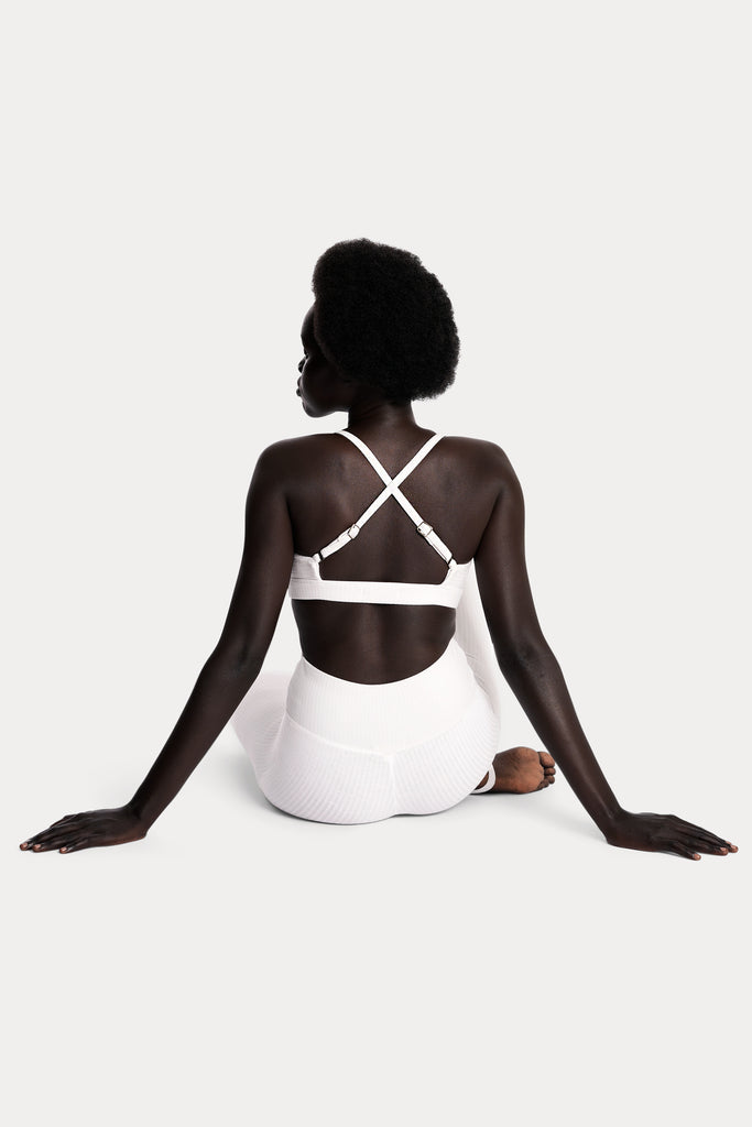 Model wearing a white ribbed bralette and matching white stirrup leggings, full-body back-side sitting on the floor.
