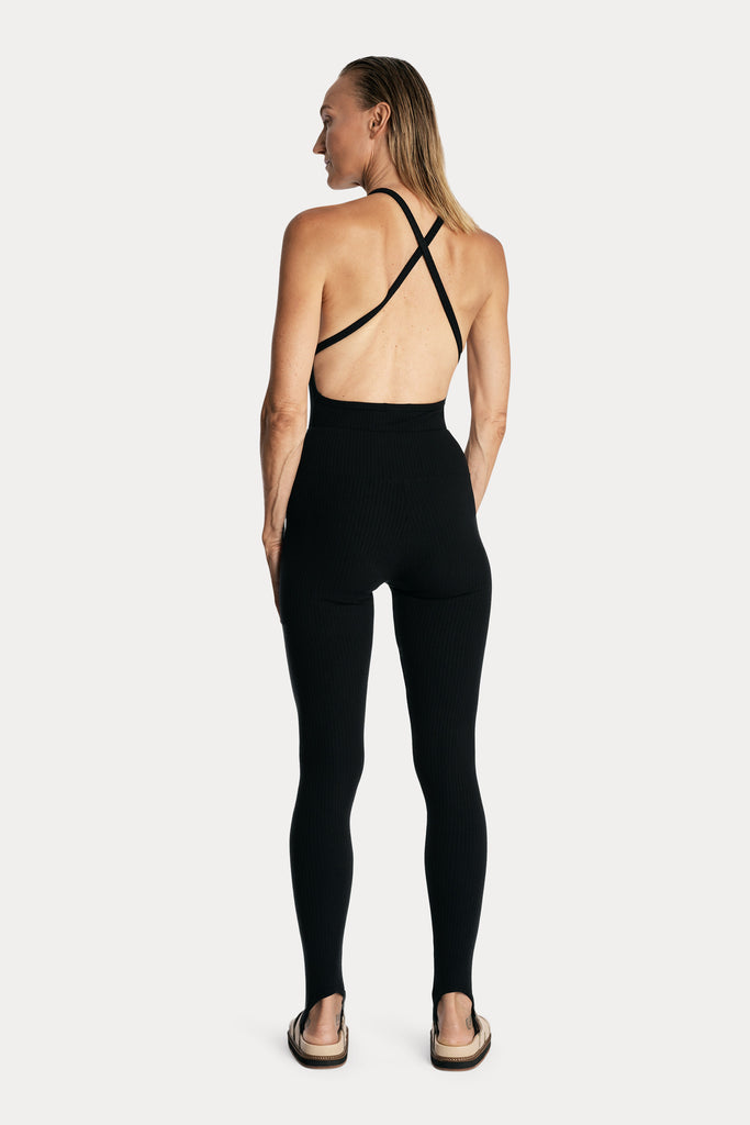 Model wearing black sustainable rib stirrup leggings and matching bodysuit, full-body back-side view.