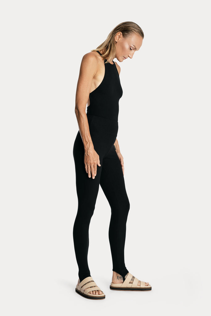 Model wearing black ribbed stirrup leggings and a matching black bodysuit, full-body right-side view.