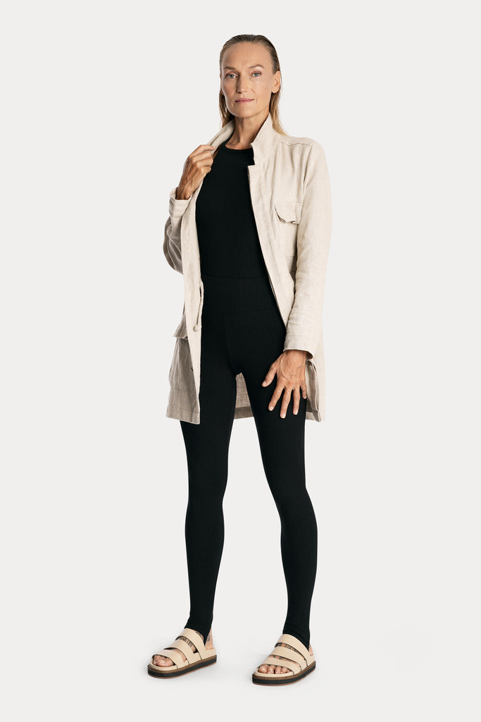 Model wearing black sustainable rib stirrup leggings and matching bodysuit, with natural linen jacket, full-body front-side view. 
