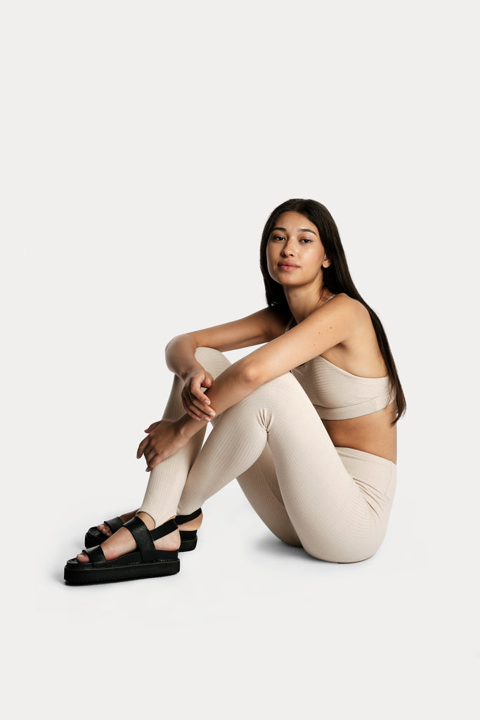 Model wearing sand ribbed stirrup leggings and a matching sand bralette, full-body left-side view while sitting on the floor.