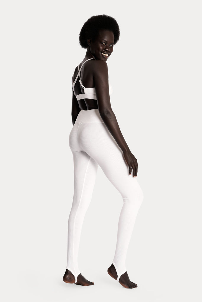 Model wearing white ribbed stirrup leggings and a matching white bralette, full-body back-right view.