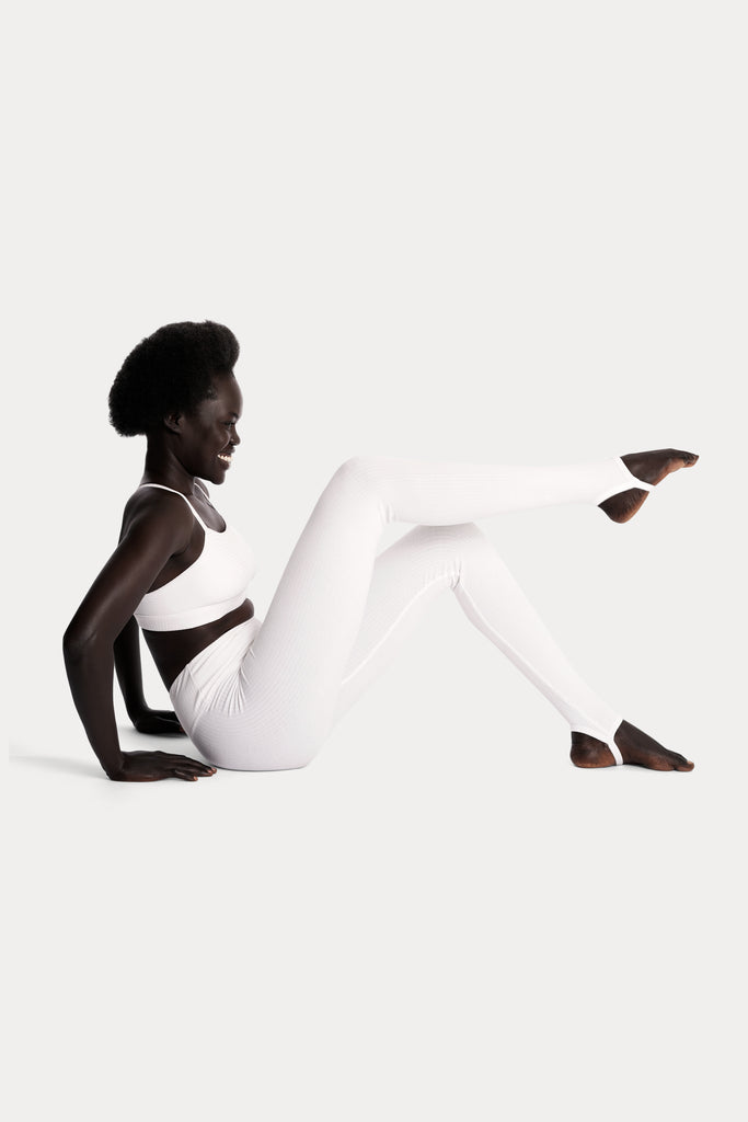 Model wearing white ribbed stirrup leggings and a matching white bralette, full-body right-side view while sitting on the floor.
