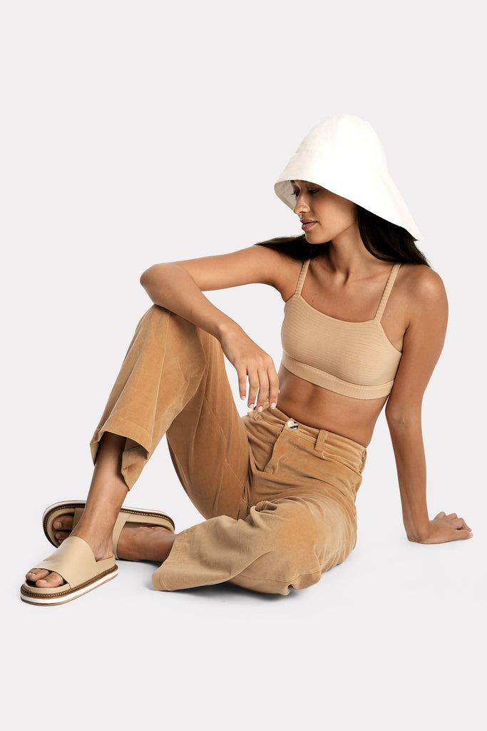 Model wearing a tan ribbed bralette and corduroy trousers, with white linen wide-brim hat, full-body front-side sitting view.